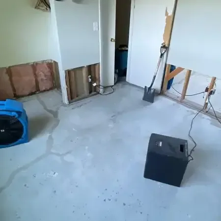 A blue air mover and a black dehumidifier on a bare concrete floor beside walls opened at the bottom for drying